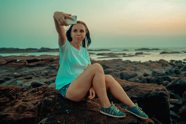 Belle fille prend une selfie sur un téléphone intelligent assise sur la plage contre le ciel du soir — Photo