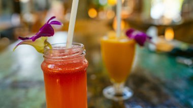 Bir tropikal kafede, arka planda portakal suyu olan sağlıklı kırmızı bir smoothie. Masada pipetli ve dekoratif çiçeklerle serin taze meyve suları ya da kokteyller..