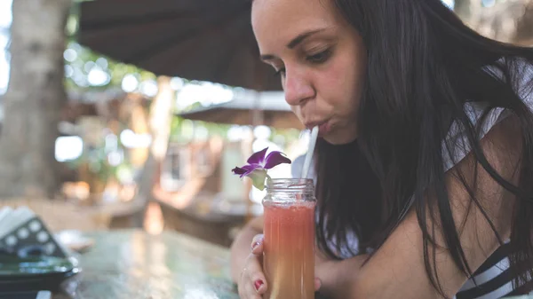 Uma jovem mulher bonita bebe um smoothie laranja saudável em um café tropical.A menina bonita bebe um suco fresco ou cocktail. — Fotografia de Stock