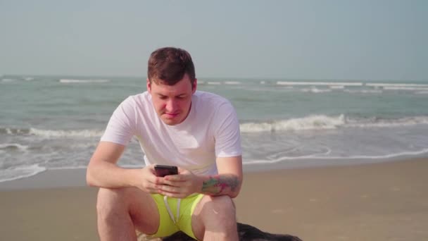 海滩上英俊的男人用他的手机。在海滩上手持智能手机的年轻人 — 图库视频影像