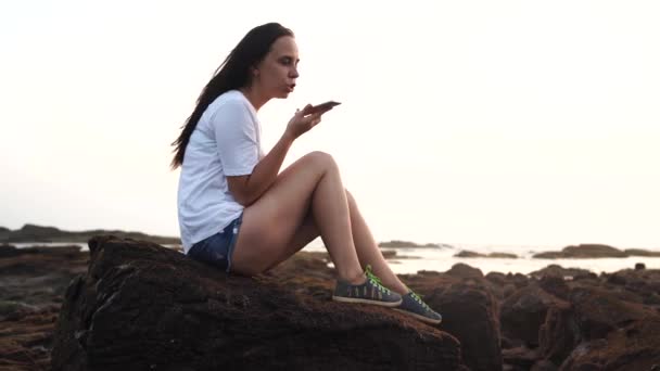 En vacker ung kvinna sitter på klipporna och spelar in ljudmeddelanden i ett socialt nätverk på en mobiltelefon vid havet mot solnedgången. — Stockvideo