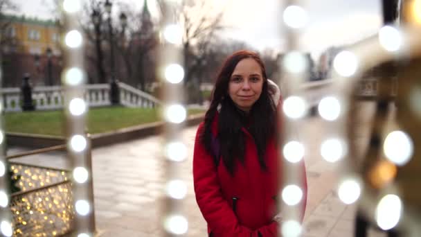 Portrait of a beautiful young woman in a jacket and hood through a garland on the street. — Stock Video