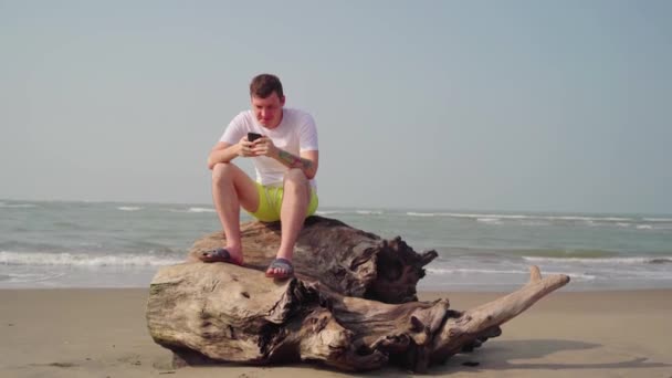 Un beau jeune homme est assis sur une bûche et feuillette les différentes nouvelles dans un téléphone portable au bord de la mer ou de l'océan par une journée ensoleillée . — Video