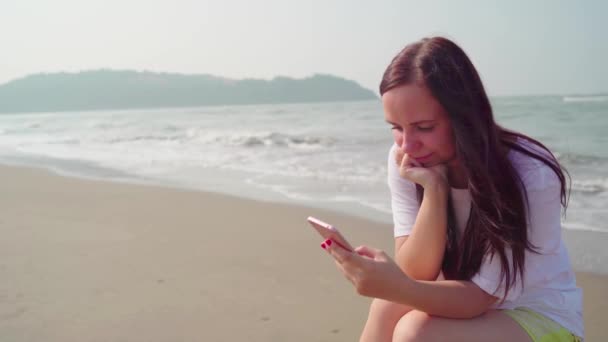 美しい若い女性が丸太の上に座って、明るい晴れた日に海や海のそばの携帯電話でさまざまなニュースを通り抜けています. — ストック動画