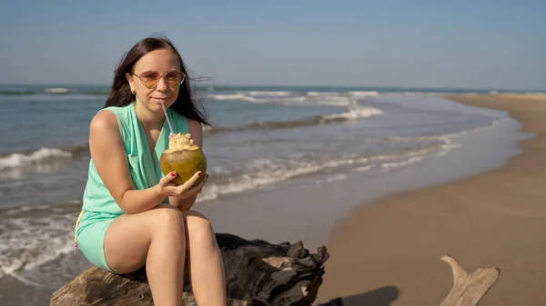 Une belle jeune femme est assise sur une bûche et boit la noix de coco au bord de la mer ou de l'océan par une journée ensoleillée. — Photo