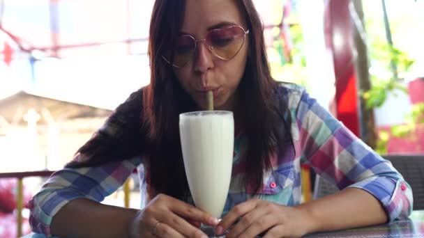 美しい若い女性が熱帯のカフェで健康的なスムージーを飲んでいます。女の子はミルクセーキを飲んでいる. — ストック動画
