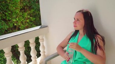 Güzel genç bir kadın sandalyede oturuyor ve terasta bir şişeden soğuk su içiyor..