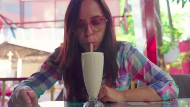 美しい若い女性が熱帯のカフェで健康的なスムージーを飲んでいます。女の子はミルクセーキを飲んでいる. — ストック動画
