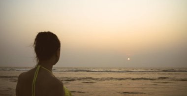 Gün batımında deniz kenarında isimsiz bir kadın. Akşamları sahilde el sallayan, mayo giymiş, tanınmamış bir kadının arka görüntüsü.