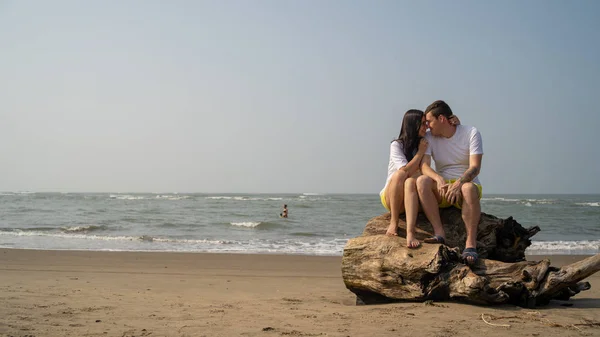 Happy couple sitting on driftwood near sea. Loving couple hugging and touching noses while sitting on driftwood during date on beach against waving sea and cloudless sky — Stock Photo, Image