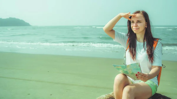 Feminino mapa de leitura turística perto do mar. Mulher descalça com mochila sentada na madeira à deriva e examinando mapa enquanto descansa na praia de areia perto do mar ondulado — Fotografia de Stock