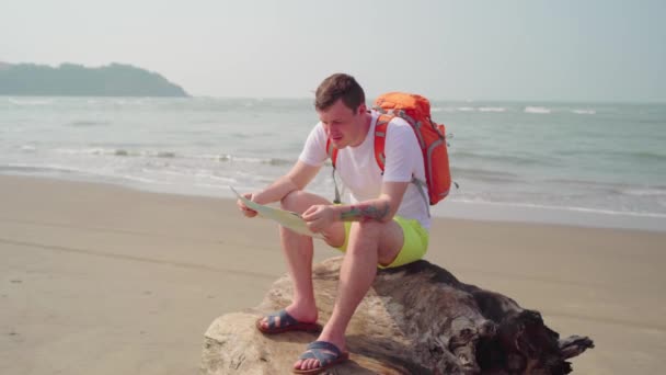 Мужской турист читает карту у моря. Взрослый мужчина с рюкзаком сидит на суше и изучает карту, отдыхая на песчаном пляже у моря. — стоковое видео