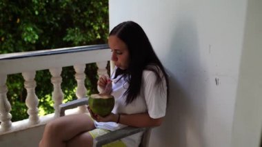 Terasta tropikal içkiden hoşlanan bir kadın. Yetişkin bir kadın balkonda sandalyede oturuyor ve güneşli bir günde dinlenirken samandan hindistan cevizi suyu yudumluyor.