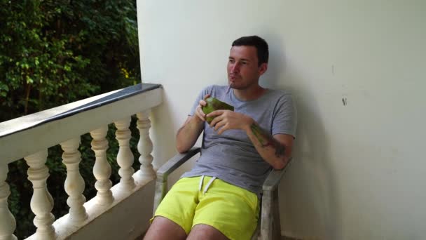 Homem que desfruta de bebida tropical no terraço. Adulto masculino sentado na cadeira na varanda e beber água de coco através de palha, enquanto descansa no resort no dia ensolarado — Vídeo de Stock
