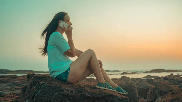 Mulher relaxada sentada na pedra e falando no smartphone na costa. Vista lateral da mulher agradável passar tempo desfrutando de férias e falando no telefone celular na costa pedregosa — Fotografia de Stock