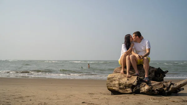 Casal feliz sentado no driftwood perto do mar. Amar casal abraçando e tocando narizes enquanto sentado no driftwood durante a data na praia contra o mar acenando e céu sem nuvens — Fotografia de Stock