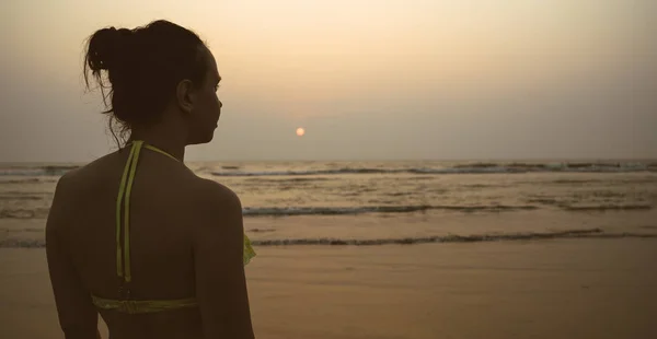 Mujer anónima cerca del mar durante la puesta de sol. Vista trasera de la mujer irreconocible con traje de baño en la playa cerca del mar agitado por la noche en el complejo. — Foto de Stock