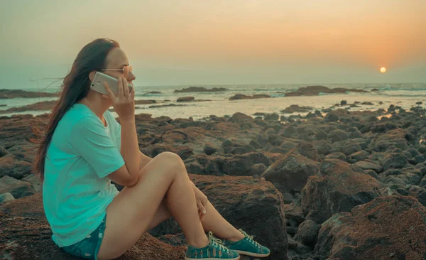 Mulher relaxada sentada na pedra e falando no smartphone na costa. Vista lateral da mulher agradável passar tempo desfrutando de férias e falando no telefone celular na costa pedregosa — Fotografia de Stock