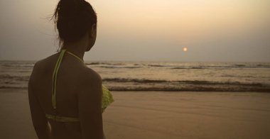 Gün batımında deniz kenarında isimsiz bir kadın. Akşamları sahilde el sallayan, mayo giymiş, tanınmamış bir kadının arka görüntüsü.