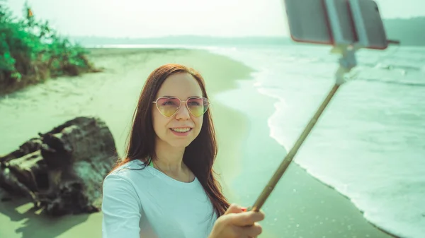 Feliz mujer tomando selfie cerca del mar. De arriba mujer feliz que utiliza monopodo con teléfono inteligente para tomar selfie cerca de mar ondulando en la playa. — Foto de Stock