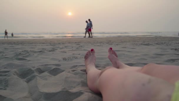 Pés femininos com pedicure vermelho encontra-se em uma praia de areia junto ao mar contra o pôr do sol . — Vídeo de Stock