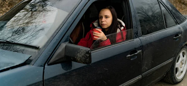 Woman with coffee in car looking out from window. Mujer de pelo largo con taza de desecho en la parte delantera del coche mirando hacia la ventana. —  Fotos de Stock