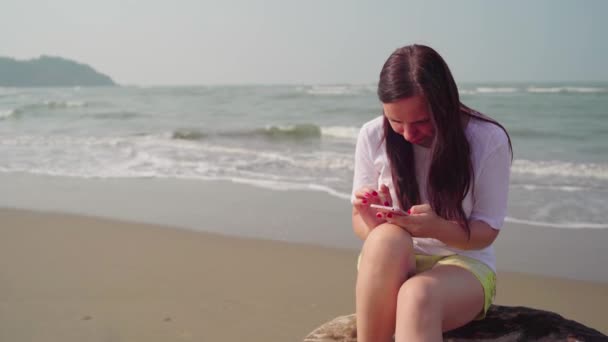 Eine schöne junge Frau sitzt auf einem Baumstamm und blättert an einem sonnigen Tag in einem Mobiltelefon am Meer oder Meer durch die verschiedenen Nachrichten. — Stockvideo