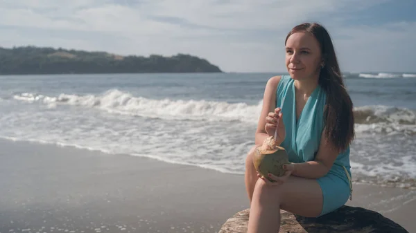 Una hermosa joven está sentada en un tronco y bebiendo el coco junto al mar o al océano en un día soleado . — Foto de Stock