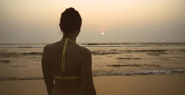 Femme anonyme près de la mer au coucher du soleil. Back view of unrecognizable female in swimwear standing on beach near waving sea in evening on resort — Photo