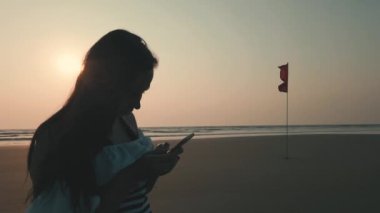 Ayakta duran ve kıyıda akıllı telefondan mesaj bırakan rahat bir kadın. Hoş bir bayanın tatilden ve deniz kenarında gün batımına karşı cep telefonuyla mesajlaşmaktan keyif almasının yan görüntüsü..