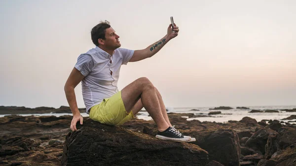 Homem relaxado sentado em pedra e tirando selfie no smartphone em terra. Vista lateral do homem agradável passar o tempo desfrutando de férias e fotografar de si mesmo no telefone celular na costa pedregosa . — Fotografia de Stock