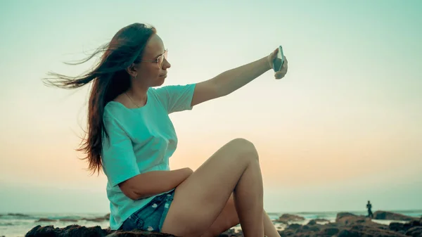 A beautiful young woman is taking selfie on a mobile phone, sitting on the rocks by the sea against the sunset. — Stock Photo, Image