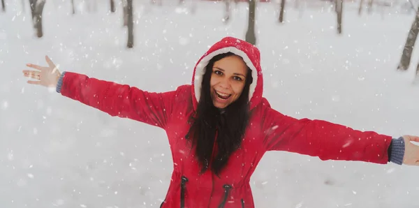 冬の赤いジャケットとフードの若い女性の肖像画。美しい女性は手を振って雪の秋に喜びます。ふわふわの雪に包まれて. — ストック写真