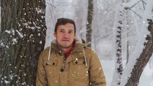 Retrato del joven en chaqueta durante la temporada invernal. El hombre guapo está cerca del árbol y sonríe coquetemente. La nieve fluida lo rodea todo.. — Foto de Stock
