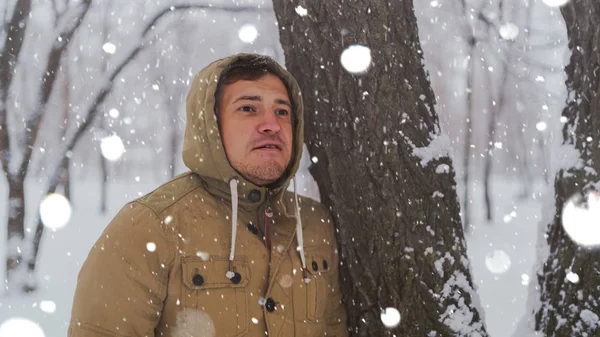 Retrato del joven en chaqueta durante la temporada invernal. El hombre guapo está cerca del árbol y sonríe coquetemente. La nieve fluida lo rodea todo.. —  Fotos de Stock