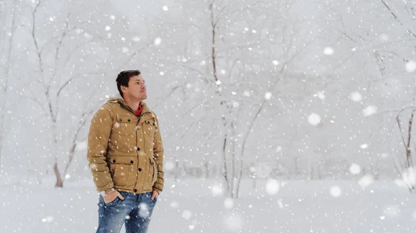 Portrait of young man in jacket and jeans in winter season. Handsome man stands and squint in winter park. Fluffy snow envelops everything around. — Stock Photo, Image