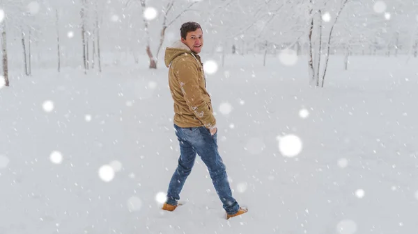 Retrato de jóvenes vestidos de chaqueta y vaqueros en temporada invernal. Un hombre agradable se pone de pie y escuche en el parque de invierno. La nieve fluida lo rodea todo.. —  Fotos de Stock