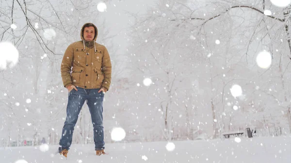 Portrait of young man in jacket and jeans in winter season. Handsome man stands and squint in winter park. Fluffy snow envelops everything around. — Stock Photo, Image