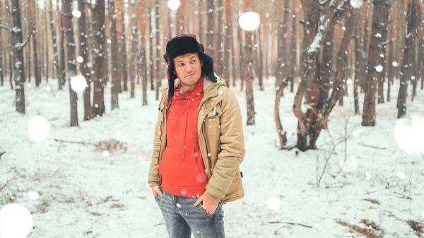 Portrait of young man in jacket and jeans in winter season. Handsome man stands and squint in winter park. Fluffy snow envelops everything around. — Stock Photo, Image