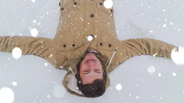 Retrato de los jóvenes en chaqueta y vaqueros, acostados en la nieve. El hombre guapo miente, extendiendo las manos y cerrando los ojos del placer en el parque invernal. La nieve fluida lo rodea todo.. —  Fotos de Stock