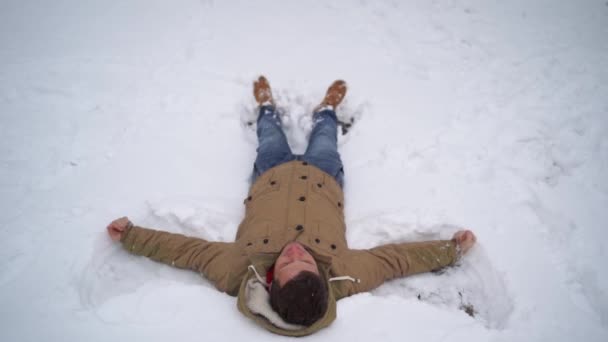 穿着夹克和牛仔裤躺在雪地里的年轻人的画像。英俊的男人躺在公园里，张开双手，闭上了眼睛，不让冬天的公园里充满欢乐。毛茸茸的雪包住了周围的一切. — 图库视频影像