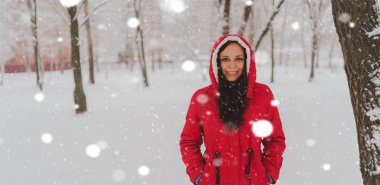 Kış mevsiminde kırmızı ceketli ve başlıklı genç bir kadının portresi. Güzel bayan gülümsüyor, karın düşüşünden zevk alıyor. Yumuşak kar her yeri kaplar..