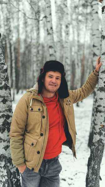 Hombre de pie en el bosque en invierno. Hombre joven y serio que lleva chaqueta de beige sobre la mandíbula roja y el sombrero de arce en el bosque nevado y mira hacia otro lado. — Foto de Stock