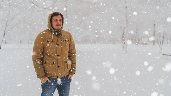 Portrait of young man in jacket and jeans in winter season. Handsome man stands and squint in winter park. Fluffy snow envelops everything around. — Stock Photo, Image