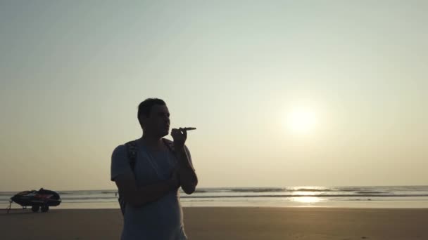 Handsome man standing and recording audio message on smartphone on shore. Portrait of pleasant man spending time enjoying vacation and talking on mobile phone on seashore against sunset. — Stock Video