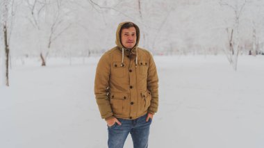 Kışın ceketli ve kot pantolonlu bir gencin portresi. Yakışıklı adam kış parkında duruyor ve gözlerini kısıyor. Yumuşak kar her yeri kaplar..