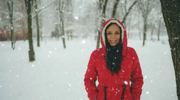 Retrato de jovem mulher em casaco vermelho e capuz na temporada de inverno. Linda senhora sorri, regozijando-se no outono de neve. Neve fofa envolve tudo ao redor . — Fotografia de Stock