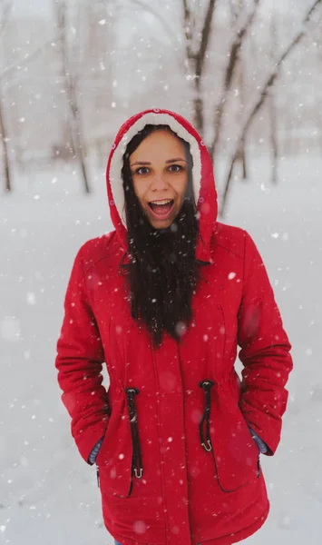 冬の赤いジャケットとフードの若い女性の肖像画。美しい女性は、雪の秋に楽しんで笑顔。ふわふわの雪に包まれて. — ストック写真