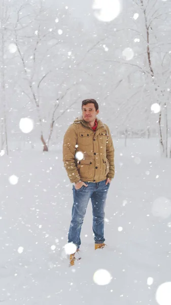 Retrato do jovem em jaqueta e jeans na temporada de inverno. Handsome homem está de pé e agachado no parque de inverno. Neve fofo envolve tudo ao redor. — Fotografia de Stock