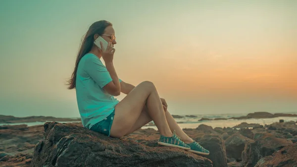 Χαλαρή γυναίκα που κάθεται πάνω σε πέτρα και μιλάει σε smartphone στην ακτή. Πλευρική άποψη της ευχάριστης γυναίκας που ξοδεύει χρόνο απολαμβάνοντας τις διακοπές και μιλώντας στο κινητό τηλέφωνο στην πετρώδη ακτή — Φωτογραφία Αρχείου